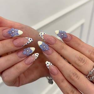 Può includere: Unghie a mandorla con stile french manicure, punte bianche decorate con pois blu. Diverse unghie presentano decorazioni floreali tridimensionali color azzurro, ognuna con al centro un piccolo strass trasparente e scintillante.