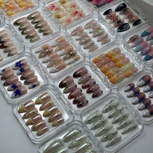 Op de afbeelding: Een verzameling van meerdere doorzichtige plastic doosjes met sets amandelvormige plaknagels. Elke set bevat verschillende ontwerpen, waaronder metallic afwerkingen, bloemmotieven, sterren, dierenprints en parelversieringen.