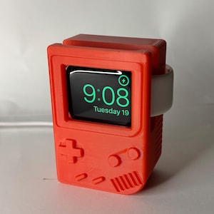 May include: A red 3D-printed Apple Watch stand in the style of a retro handheld gaming console. The watch face shows the time, 9:08, and the date, Tuesday 19. The stand features a cross-shaped D-pad and other gaming-themed buttons.