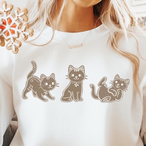 Puede incluir: Sudadera blanca con tres ilustraciones de gatos marrones. Los gatos están en diferentes poses, con detalles decorativos. La sudadera tiene cuello redondo y mangas largas. Una galleta de jengibre es visible en la esquina superior izquierda.