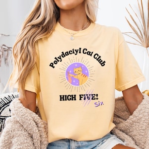 May include: Yellow t-shirt with the text "Polydactyl Cat Club" and a graphic of a cat giving a high five. The graphic is inside a purple circle with sun rays. The words "HIGH FIVE! Six" are below the graphic.