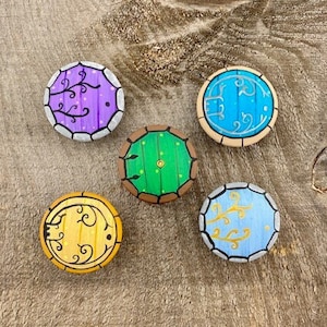 May include: Five circular wooden door decorations in various colours. Each door is painted with a different colour: purple, green, blue, yellow, and light blue. The doors have decorative swirls and are set on a wooden surface.