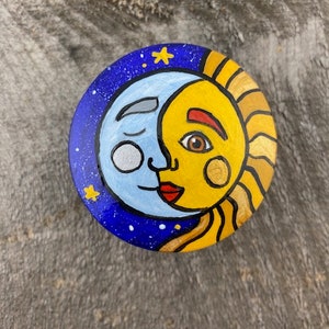 Puede incluir: Un diseño celestial redondo pintado a mano que representa un sol y una luna sonrientes. La luna es azul claro con un ojo cerrado, y el sol es amarillo con un ojo marrón. El fondo es azul oscuro con estrellas blancas y detalles dorados.