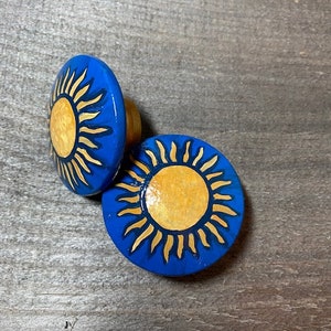May include: Two blue and gold sun-shaped wooden knobs. The knobs have a raised sun design with a yellow center and gold rays.