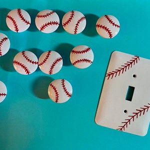 May include: Nine white baseball-themed knobs with red stitching and a white light switch cover with red stitching. The knobs and switch cover are on a turquoise background.