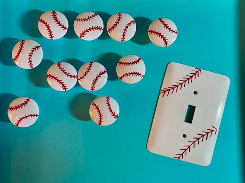 May include: Nine white baseball-themed knobs with red stitching and a white light switch cover with red stitching. The knobs and switch cover are on a turquoise background.