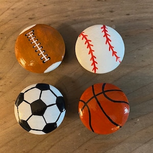 May include: Four painted rocks, each depicting a different sport: a brown and white football, a white and red baseball, a black and white soccer ball, and an orange and black basketball.