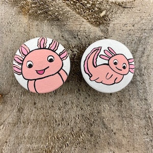 Peut inclure: Deux cercles en bois peints avec des motifs d'axolotl roses et blancs. Les axolotls sourient et ont de petits yeux noirs.