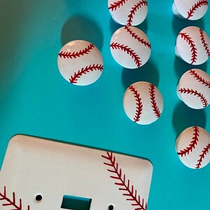 May include: White light switch cover with a baseball design. The cover has red stitching and is surrounded by baseball-themed knobs.