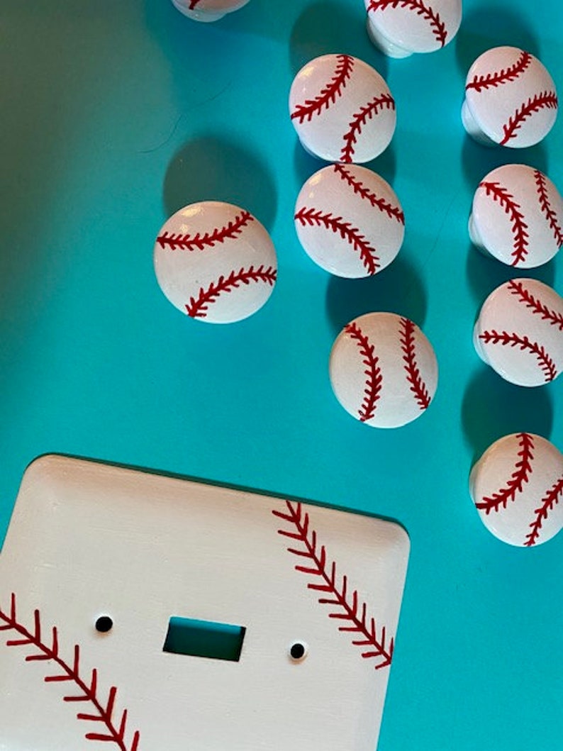 May include: White light switch cover with a baseball design. The cover has red stitching and is surrounded by baseball-themed knobs.