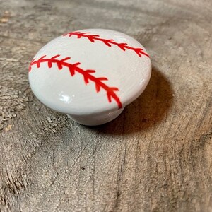 May include: White ceramic knob with a red baseball design. The knob is round and has a raised design of a baseball with red stitching.