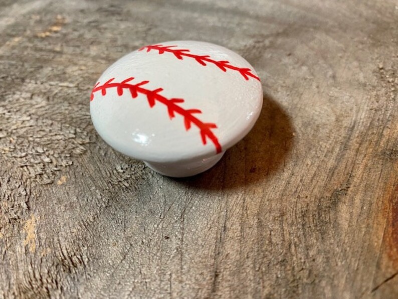 May include: White ceramic knob with a red baseball design. The knob is round and has a raised design of a baseball with red stitching.