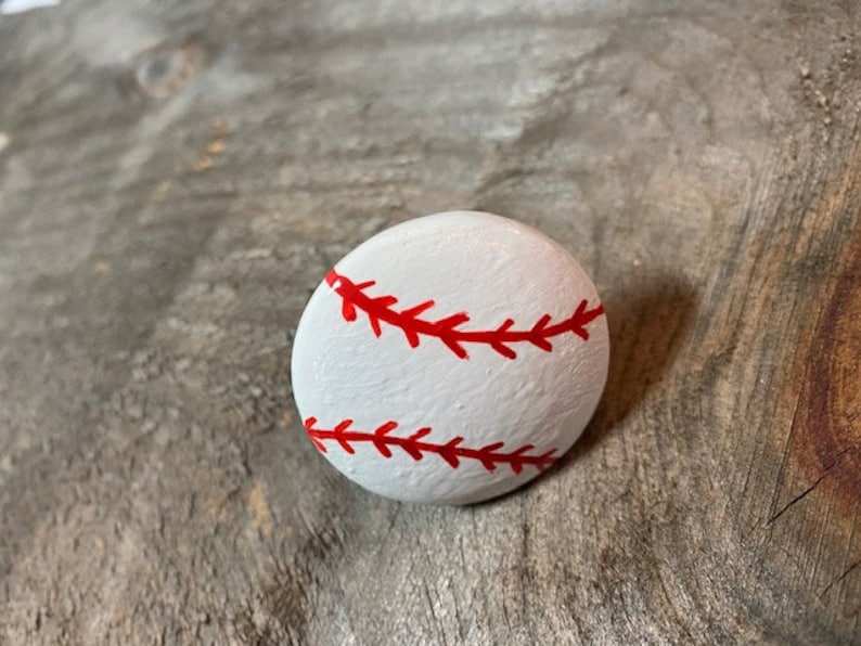 May include: A white painted rock with red stitching, resembling a baseball.