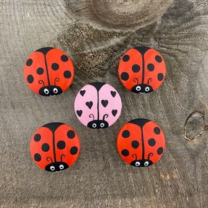 Puede incluir: Cinco piedras de mariquita pintadas a mano. Cuatro son rojas con lunares negros y cabeza negra. La quinta es rosa con corazones negros y cabeza negra. Las piedras están dispuestas sobre una superficie de madera.