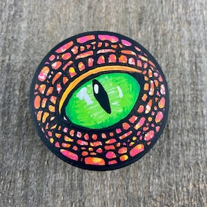 Peut inclure: Une pierre ronde peinte en noir, ornée d'un œil vert vif avec une pupille noire en forme de fente. L'œil est entouré d'un motif détaillé d'écailles orange, roses et jaunes, créant un design artistique saisissant. Sur fond de bois.