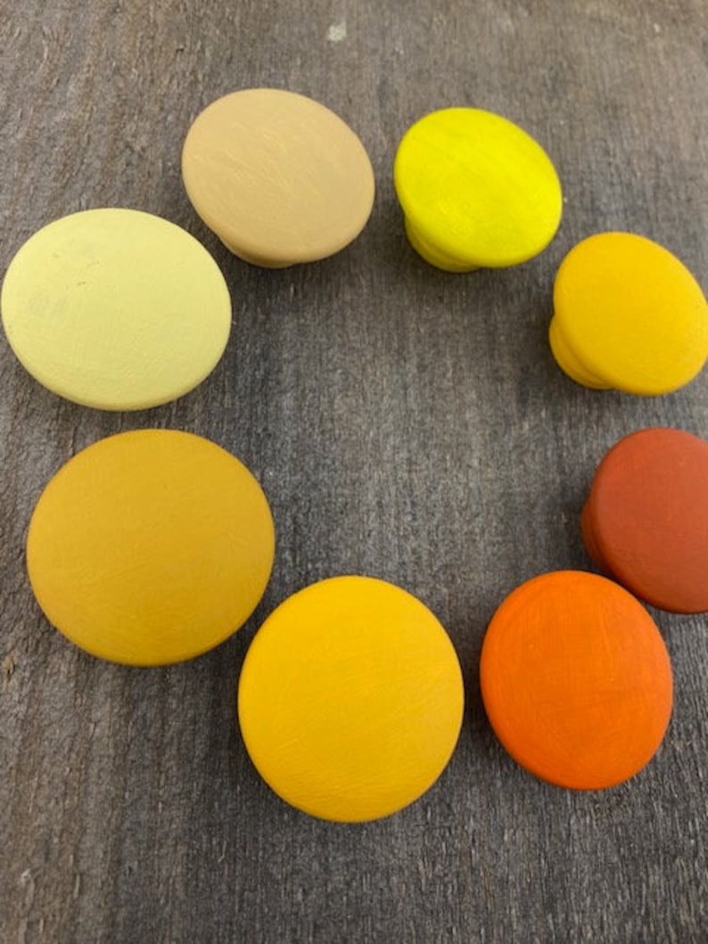 May include: Nine round wooden knobs in various shades of yellow, orange, and brown. The knobs are arranged in a circle on a wooden surface.
