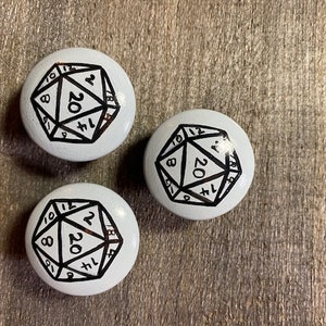 Peut inclure: Trois boutons ronds en céramique blanche avec des dessins à l'encre noire d'un dé à vingt faces. Chaque dé porte les numéros 1, 2, 3, 4, 5, 6, 7, 8, 9, 10, 11, 12, 14, 16, 18, 20.