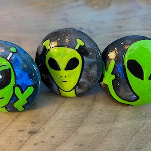 May include: Three hand-painted rocks featuring green alien faces against a starry, dark blue and black background. The aliens have large black eyes and are painted with bright green paint. The rocks are on a wooden surface.