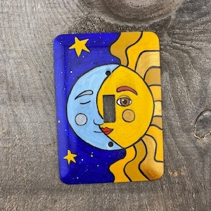 May include: Hand-painted light switch cover featuring a celestial design. The left side depicts a blue moon with closed eyes and the right side shows a yellow sun with an open eye. The background is a dark blue with yellow stars.
