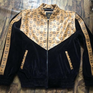 Puede incluir: Chaqueta negra con cremallera y estampado dorado con las letras "MCM" repetidas en el pecho y las mangas. La chaqueta tiene una textura negra aterciopelada y un cuello dorado. Prenda de moda.