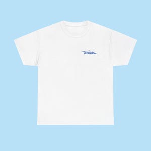 May include: White t-shirt with a crew neck and short sleeves. The shirt features the word "Iceman" in blue script on the upper left chest area. The background is a light blue.