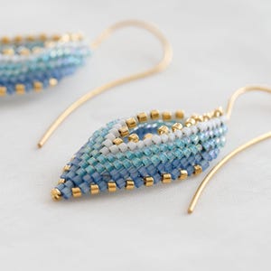 Peut inclure: Une paire de boucles d'oreilles artisanales en forme de feuille. Les boucles d'oreilles présentent un dégradé de perles bleues et blanches, rehaussées de perles dorées. Les crochets d'oreilles sont dorés. Les boucles d'oreilles sont présentées sur une surface blanche.