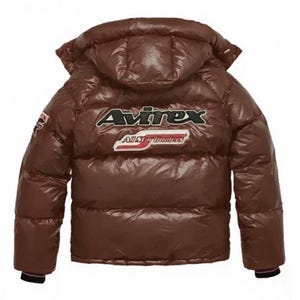Puede incluir: Chaqueta acolchada marr&oacute;n con capucha. La parte trasera de la chaqueta presenta la marca "Avirex" en letras blancas y negras, con "All Star Dealers" debajo. La chaqueta tiene una textura brillante y acolchada.