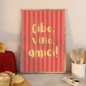 May include: A framed print with the words "Cibo, Vino, amici!" in yellow script on a pink and red striped background. The print is displayed on a kitchen counter with a red toaster, bread, and utensils.