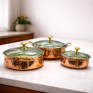 Puede incluir: Tres ollas de cocina de color cobre con tapas de vidrio y asas y perillas doradas. Las ollas están dispuestas en fila, con la olla más grande al frente y la más pequeña atrás. Las ollas tienen una textura martillada.