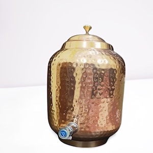 May include: A hammered copper-colored beverage dispenser with a lid and a spigot. The dispenser has a textured surface and a small, decorative knob on top. The spigot is silver-colored with a blue and white label. The dispenser sits on a round base.