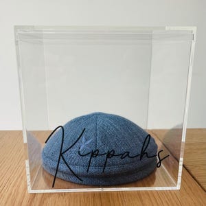 May include: A blue kippah with a star design, displayed inside a clear acrylic box. The word "Kippahs" is written in black script on the box. The kippah is made of a textured fabric and sits on a wooden surface.