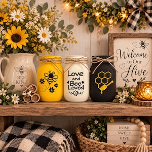 May include: Three decorative glass jars with twine bows, featuring bee-themed designs. One yellow jar with a black bee, one cream jar with the text Love and Bee Loved, and one black jar with a yellow honeycomb and dipper graphic on a wooden shelf.