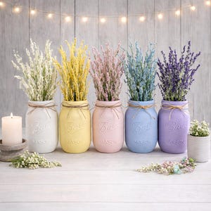 May include: Five painted glass Ball Mason jars in white, yellow, pink, light blue, and purple, each tied with twine and filled with matching colored faux floral sprigs. The jars are arranged in a row, with a white candle and small floral accents.