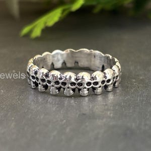 May include: A silver ring with a repeating skull pattern. The ring is made of a shiny metal and has a detailed, gothic aesthetic. The ring is a band style, with the skulls forming the entire outer surface. The text "925Jewels" is visible on the ring.