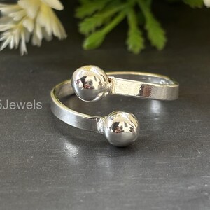 Può includere: Un anello color argento con due accenti sferici. L'anello ha un design minimalista, con una fascia che avvolge il dito e presenta una piccola sfera a ciascuna estremità. Il testo "925Jewels" è visibile.