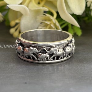 Puede incluir: Un anillo de plata con una banda de elefantes tallados. El anillo tiene un acabado pulido y presenta el texto "925Jewels" en el lateral. Los elefantes están dispuestos en un patrón repetitivo alrededor de la banda.