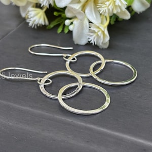 Puede incluir: Pendientes colgantes plateados con una serie de anillos martillados interconectados. Los pendientes tienen un cierre de gancho y se muestran sobre un fondo oscuro con flores blancas. El texto "925 jewels" es visible.