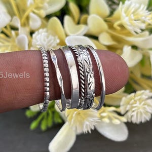 Può includere: Una collezione di anelli d'argento, ognuno con un design unico, tra cui modelli con perline, intrecciati e floreali. Gli anelli sono mostrati su un dito su uno sfondo di fiori bianchi e gialli. Il testo "925Jewels" è visibile.