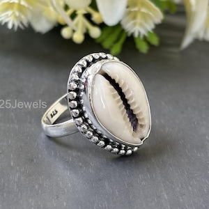 May include: A sterling silver ring featuring a large, oval-shaped cowrie shell. The shell is cream-colored with a dark central stripe, set in a beaded bezel. The ring band is simple and polished, with the text "925 Jewels".