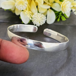 Könnte beinhalten: Ein poliertes silbernes Armreif-Armband mit geschwungenem Design. Das offene Armband wird vor einem dunklen Hintergrund mit einem verschwommenen Blumenarrangement gezeigt. Das Armband hat einen Durchmesser von ca. 6,35 cm.