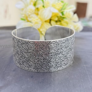 May include: A wide, silver cuff bracelet with an intricate, swirling floral pattern. The open-ended design allows for easy wear. The bracelet is approximately 2 inches wide.