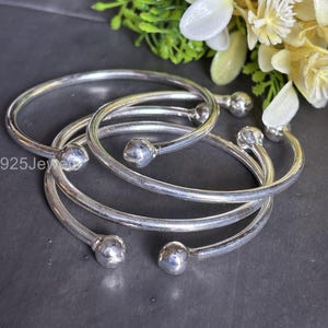 Może przedstawiać: Kolekcja srebrnych bransoletek. Każda bransoletka ma gładki, zaokrąglony kształt z małym, kulistym akcentem na każdym końcu. Bransoletki są ułożone w stos, a widoczny jest napis "925Jewelry".