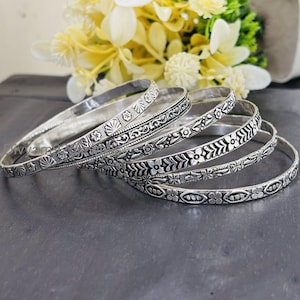 May include: A collection of silver bangles with intricate floral and geometric patterns. The bangles are arranged in a slightly overlapping manner, showcasing the detailed designs. The background features yellow and white flowers, adding a touch of color.