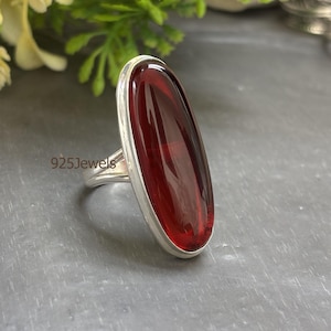 May include: A silver ring with a large, oval, deep red gemstone. The gemstone is set in a silver bezel and has a polished surface. The ring is displayed on a gray surface with flowers in the background. The text "925Jewels" is visible.