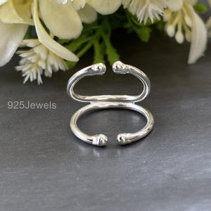Könnte beinhalten: Ein silberner Ring mit einem einzigartigen offenen Design. Der Ring hat zwei gebogene Bänder, eines bildet die Basis und das andere ein paralleles oberes Band, jedes mit abgerundeten Enden. Der Text "925Jewels" ist auf der linken Seite sichtbar.
