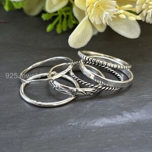 May include: A collection of silver rings in various designs. The rings include a twisted rope style, a band with small square details, and rings with floral and geometric patterns. The rings are displayed on a dark surface with a floral background.