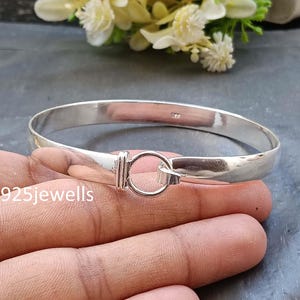 May include: A polished silver bangle bracelet with a circular clasp. The bracelet is round and smooth, with a simple, elegant design. The clasp is a small, round ring that connects the two ends of the bracelet. The bracelet is shown on a person's hand.