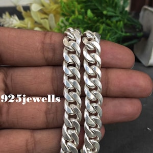 Puede incluir: Una pulsera de cadena tipo barbada en tono plateado con eslabones ovalados gruesos entrelazados y un acabado pulido y reflectante. El texto 925jewells está impreso en blanco en el lado izquierdo, cerca de los eslabones de metal.