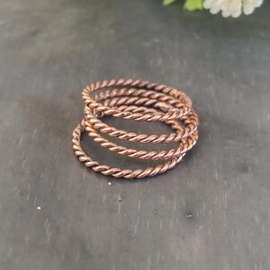 Peut inclure: Cinq bagues de couleur cuivre, torsadées en forme de corde, empilées les unes sur les autres. Les bagues sont faites d'un matériau métallique brillant. La bague du bas est légèrement plus grande que les autres.