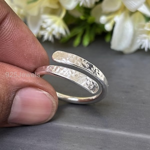 Puede incluir: Un anillo envolvente plateado con una textura martillada. El anillo tiene un diseño curvo, con los extremos superpuestos. El anillo está sostenido por los dedos de una persona. El texto "925 Jewels" es visible en el anillo.